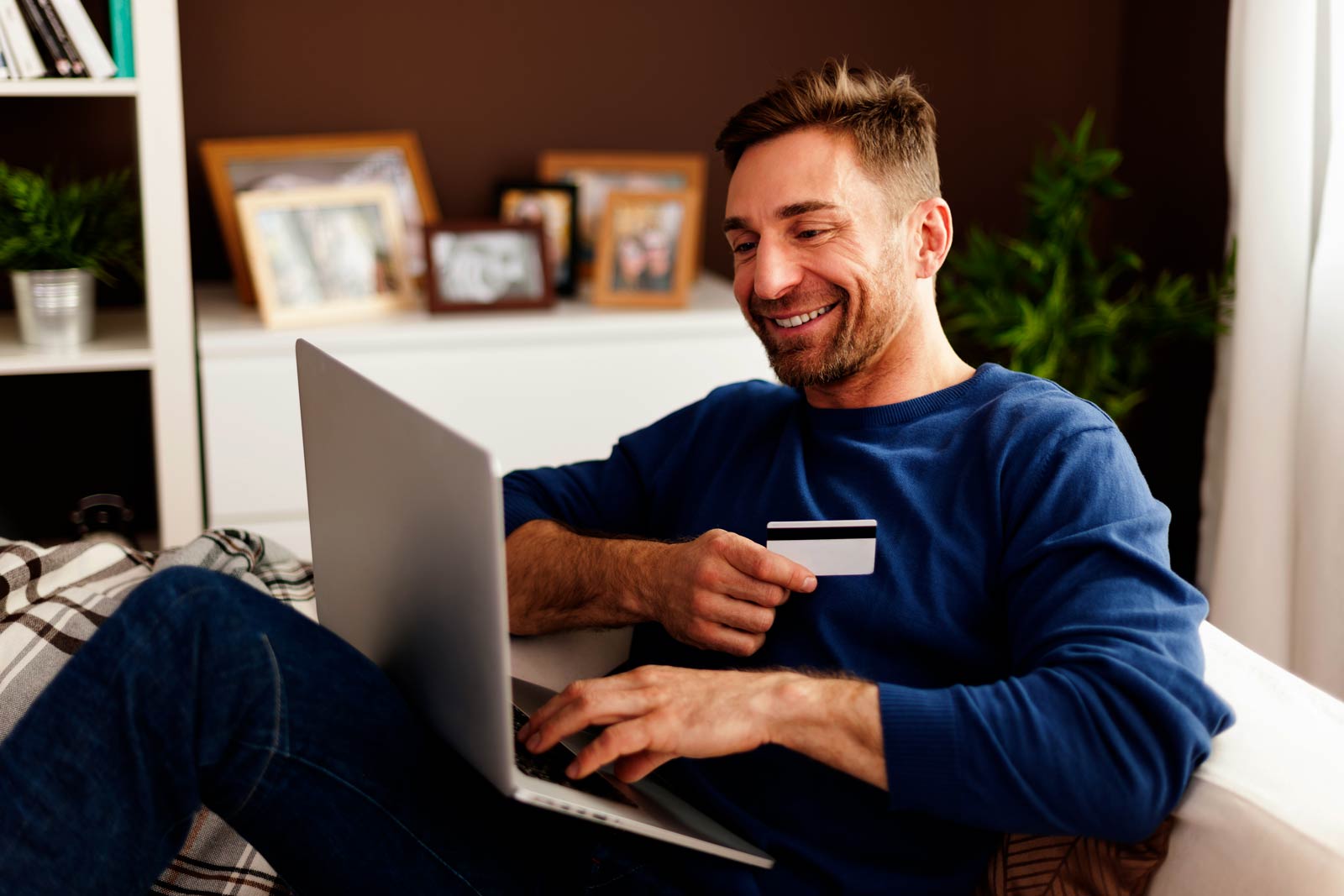 Homem sorrindo no sofá com cartão de crédito na mão e notebook no colo